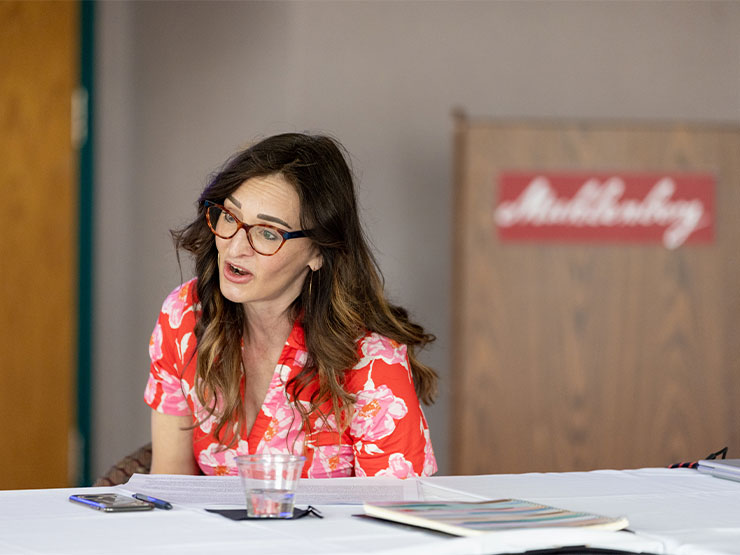 An instructor with long brown hair and glasses wears a colorful pink and white blouse while speaking across a table.