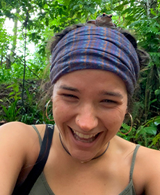 A person, wearing their hair back in a plaid bandana, laughs in a tropical forest.