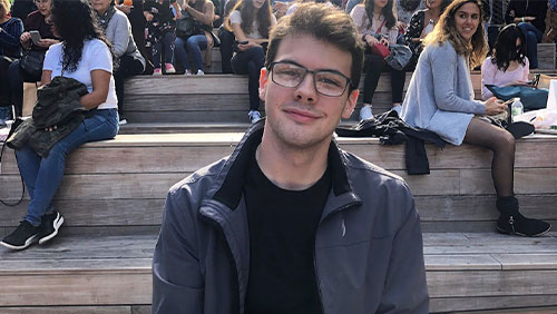 A young man with dark hair and glasses in a black shirt and jacket smiles at the camera while sitting on some bleachers