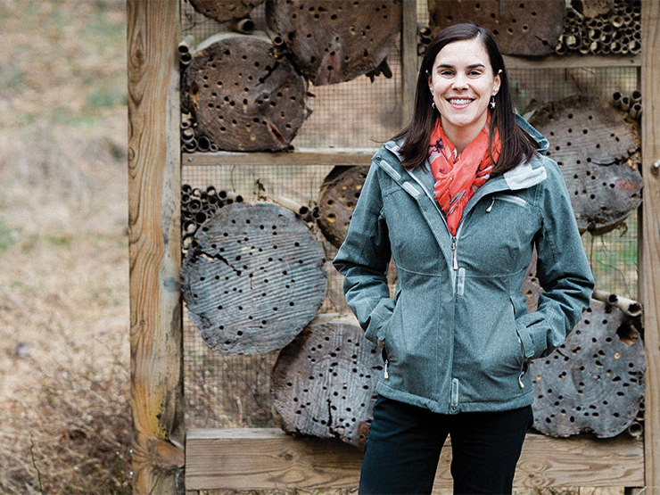 A person in a grey jacket stands outside near a structure with logs dotted with holes for bees.