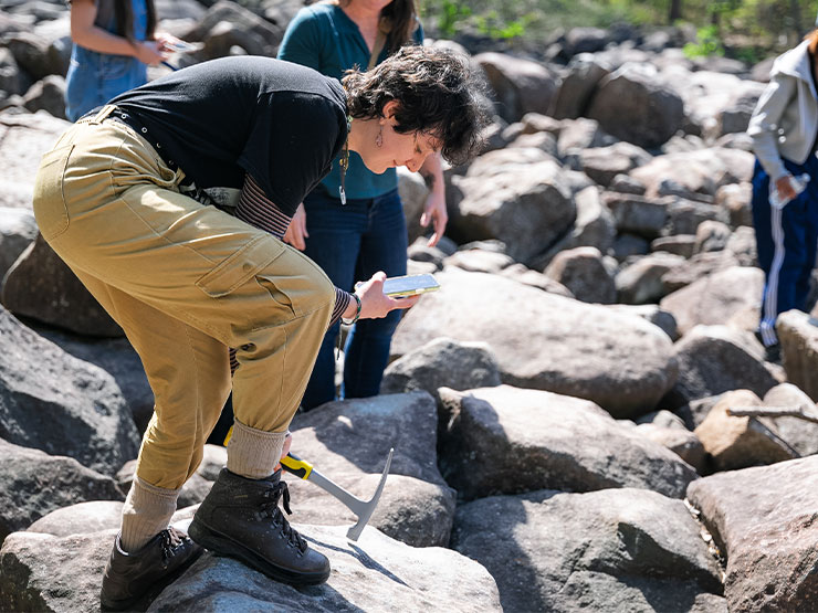 A college student leans over a rock in a boulder field, cell phone and hammer in hand.