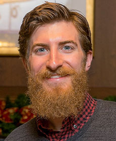 A smiling adult with a beard wears a dark grey sweater over top a red and blue checkered plaid shirt.