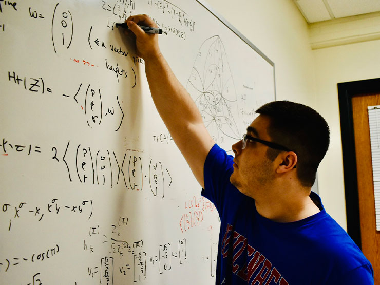 A college student reaches high on a whiteboard to add to a complicated formula.