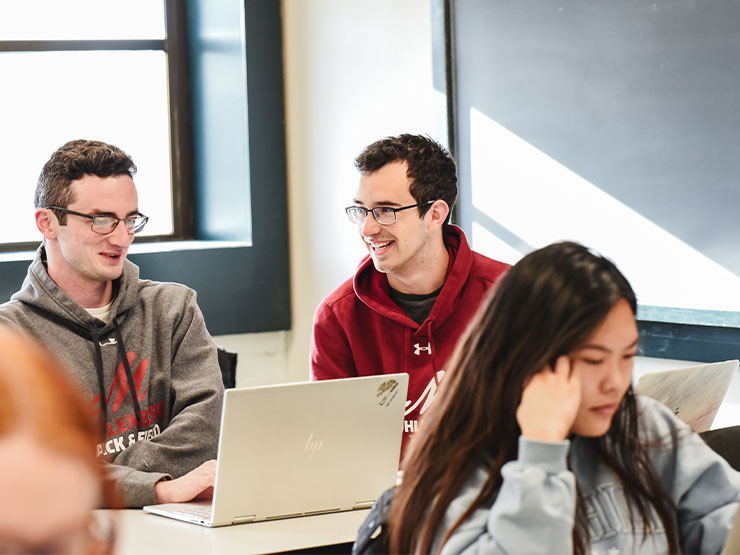 A pair of college students smile at one another while working on open laptops in a classroom.