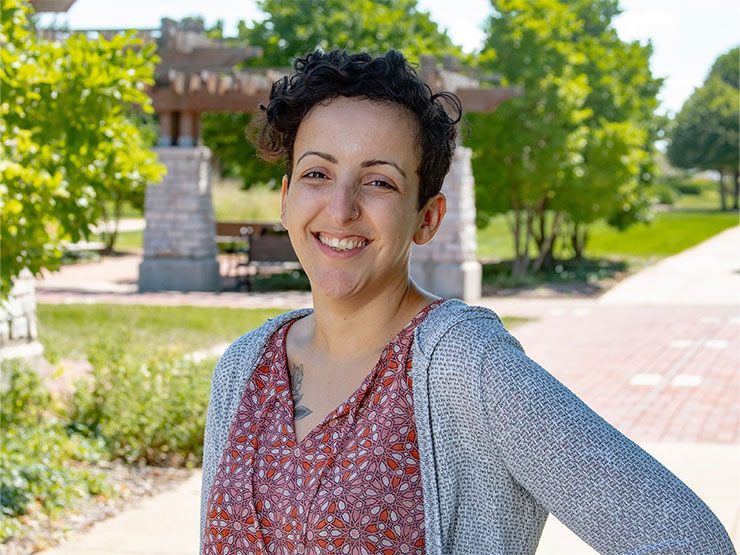 A headshot taken outside of an adult with short dark hair and is wearing a flowered top and gray sweater