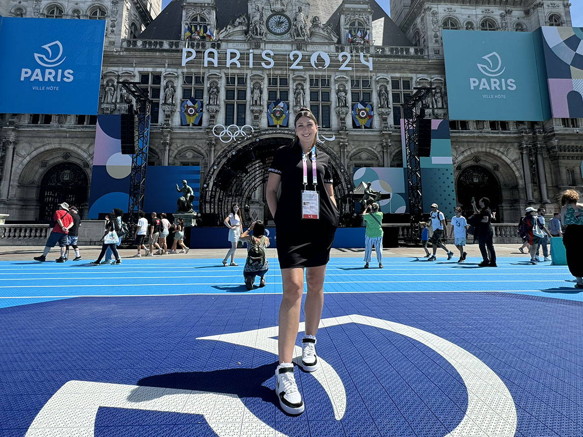 A smiling woman with a lanyard around her neck stands outside of gothic building decorated with signs for the Paris Olympics.