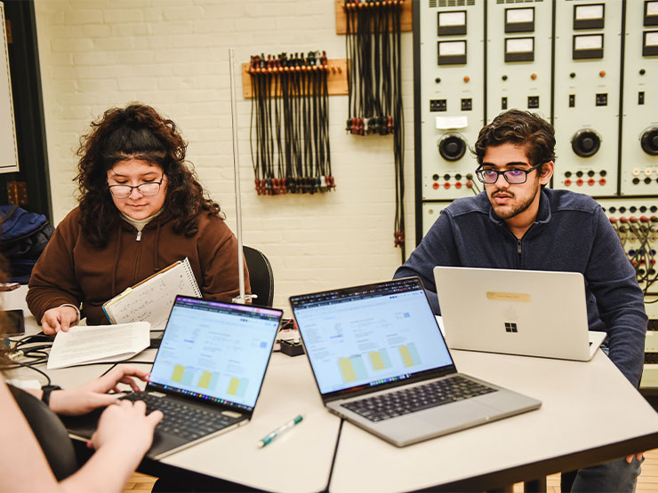 A group of students work together on laptops at a shared desk space in a lab with lab equipment hanging on a wall behind them.