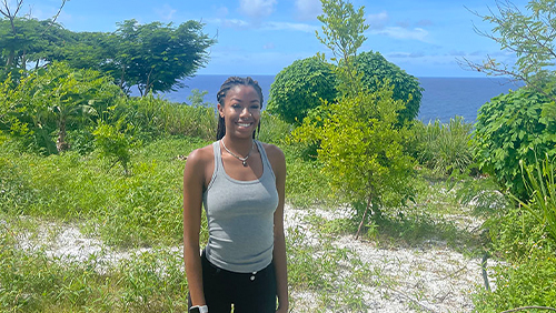 A Black woman with brown hair and athletic attire standing in a grassy field neutrally posing for a picture.