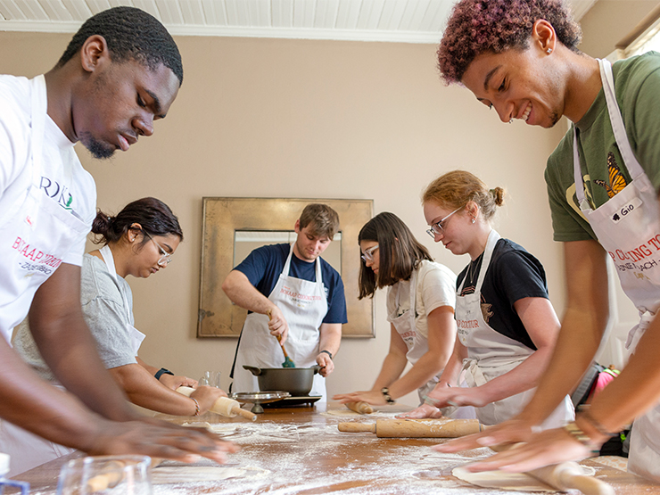 A group of students wearing white aprons knead dough on a large table scattered with flour.