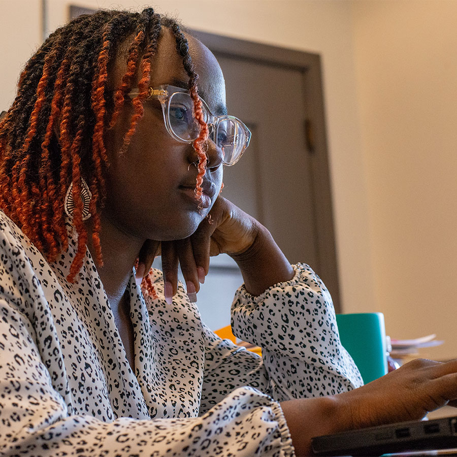 A young adult with glasses and black cornrows with orange ends sits at a desk and looks at a computer screen, face resting on their hand.