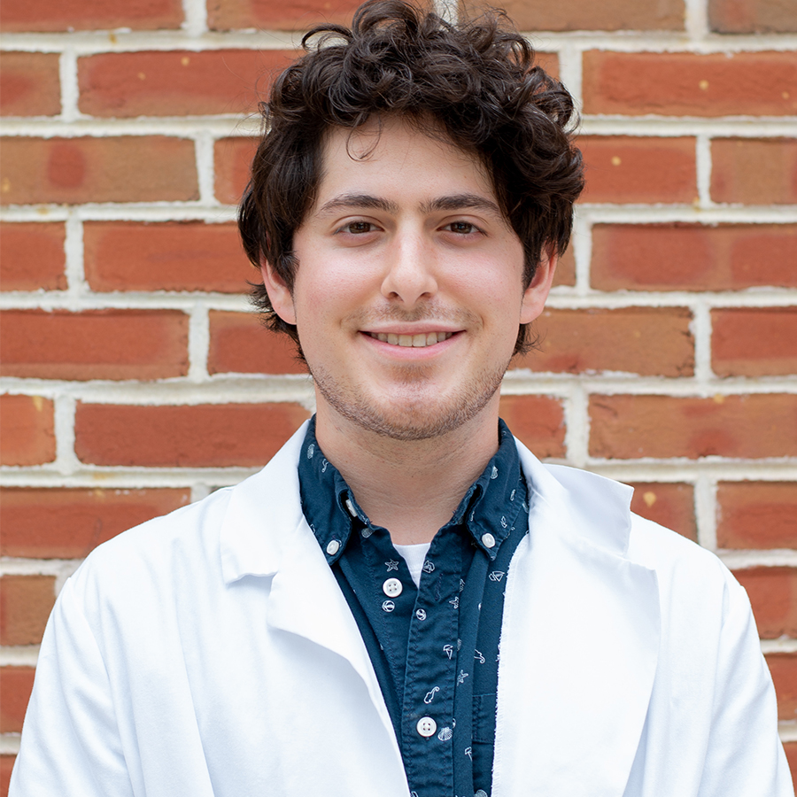 A student in a white lab coat smiles at the camera while standing in front of a brick wall.