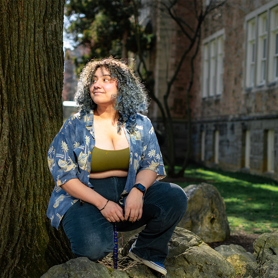 A student with curly black and blue hair kneels next to a tree on campus and looks off into the distance.