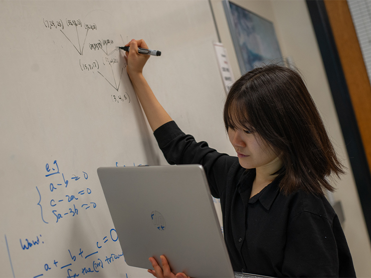 A student writes formulas on a white board with one hand while consulting an open laptop held in the other.