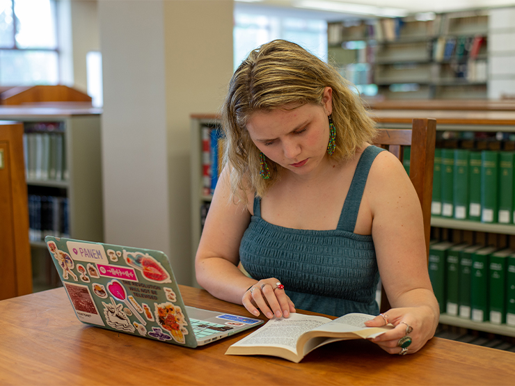 A student examines a passage in a book while conducting research at a library desk, her sticker-covered laptop open in front of her.