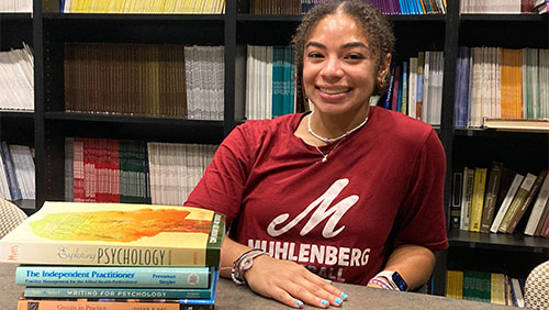 A college students smiles at a desk with a pile of psychology books nearby and a packed bookshelf behind her.