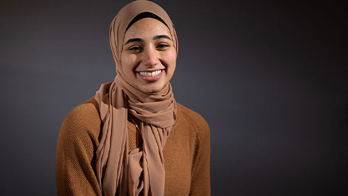 A young adult in a tan sweater and matching headscarf smiles at the camera in front of a gray background.