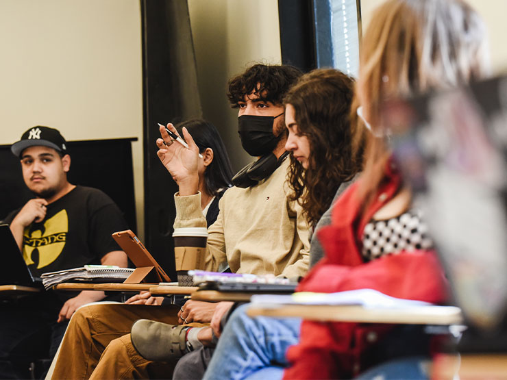 A student in a classroom, wearing a black face mask, raises a hand to speak.