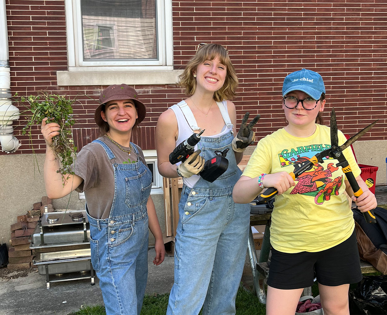 Three young students in outdoor work clothes smile and pose while holding gardening gear and plants.