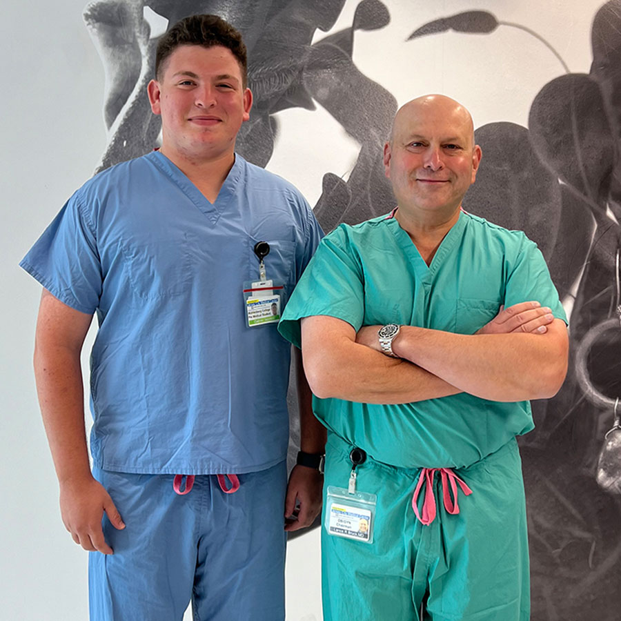 A college student in blue scrubs stands next to a doctor in green scrubs.