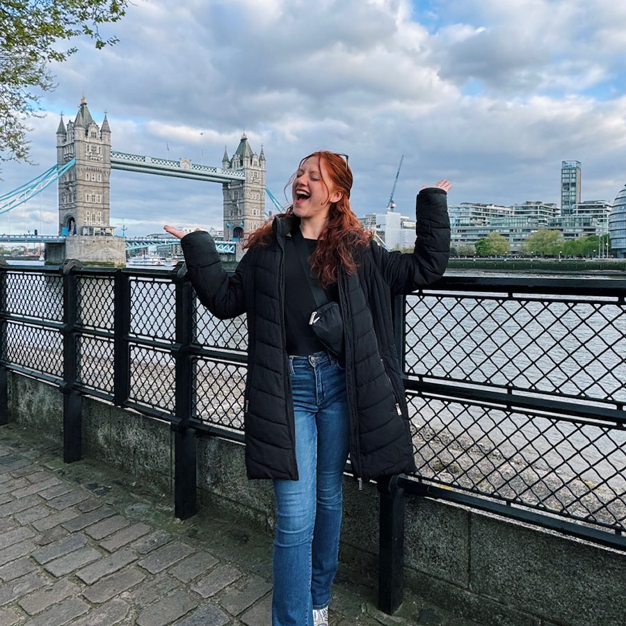 A college student with long red hair poses with her arms outstretched outdoors, alongside a river with the London Bridge in the distance.