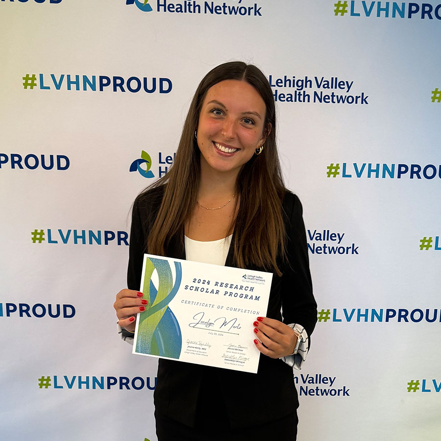 A young adult in a blazer and skirt holds a certificate in front of a background that reads 