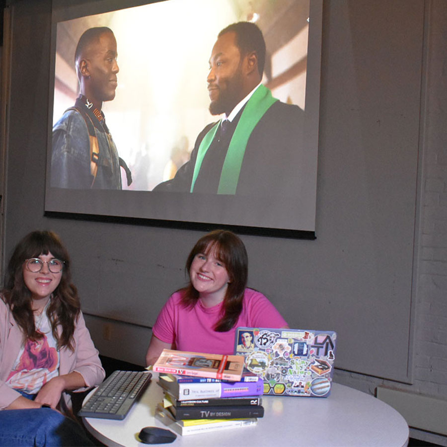 An instructor and a college student sit side-by-side at the table while a movie plays on a projector screen behind them.