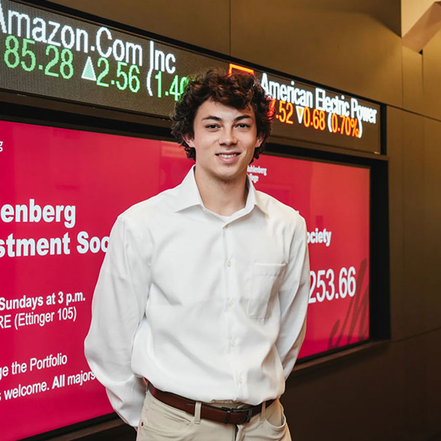 A college student stands in front of a stock ticker and video board that says Muhlenberg Investment Society