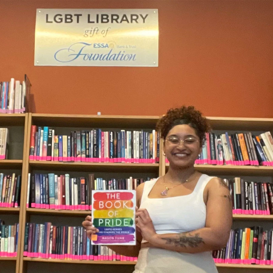 College student Princesa Santos holds a book in front of the bookshelves of an LGBT Library.