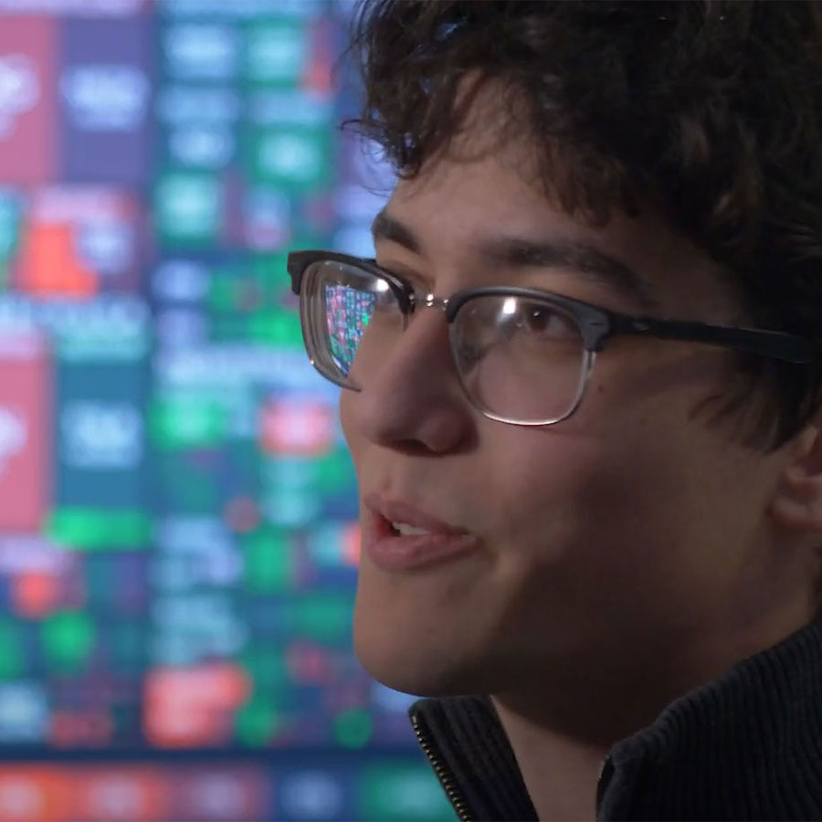 A college student with glasses speaks to someone off-camera, a screen full of stock information in the background.