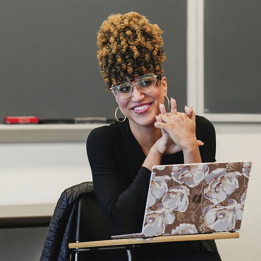 A college professor sits in front of her class with a laptop open on the desk in front of her