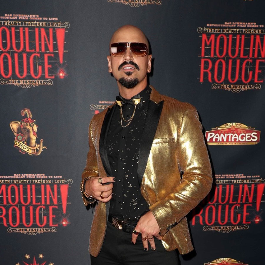 A man appears on the red carpet in a black dress shirt, golden blaze and sunglasses against a backdrop for the Broadway performance of Moulin Rouge.