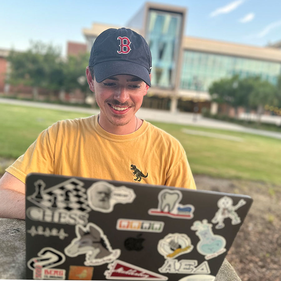 A smiling college student works on a laptop outside at a college campus.