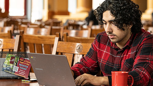 A student wearing a red flannel shirt sits in a dining hall with an open laptop and a red mug.
