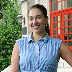 A woman in a light blue sleeveless dress stands, one arm on her hip, outdoors on the campus of Muhlenberg College.