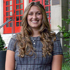 A woman with long curly hair stands in a dress outdoors, smiling on the campus of Muhlenberg College.