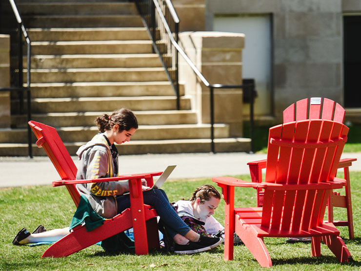 A pair of College students work outdoors, one seated in a red Adirondak chair, and another lying on the ground nearby.