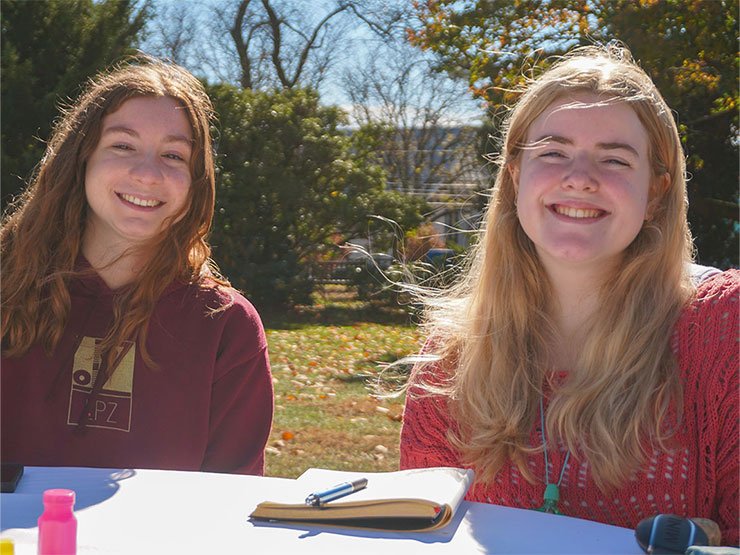 A pair of students sit smiling at a table outdoors with a notebook in front of them.