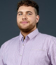 Assistant Director of Multicultural Life & Specialty Housing, Zakary Bray, wearing blue button up collar shirt in front of blue background.