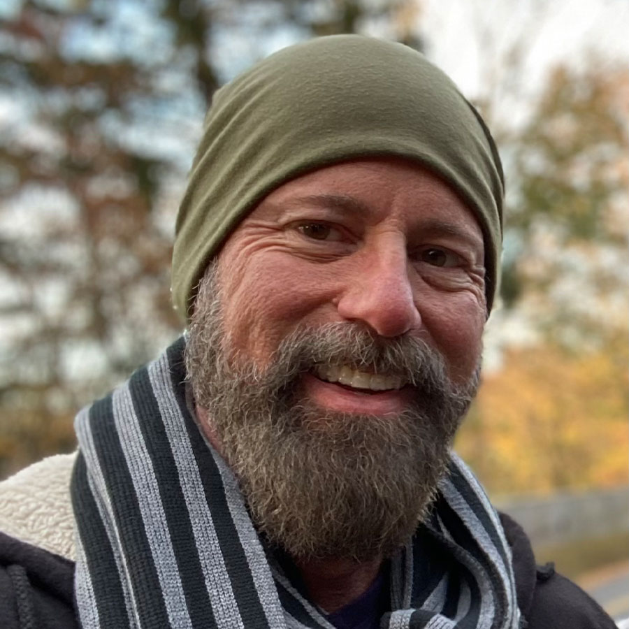 A smiling man with a beard wears a green hat and striped scarf outdoors.
