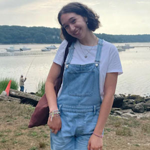 A college student in overalls smiles at the camera, head titled, while standing outdoors at the edge of a lake.