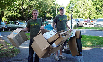 Students in green t-shirts haul cardboard boxes on a college campus.