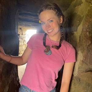 A college student in a pink t-shirt poses with a smile inside a cavern.