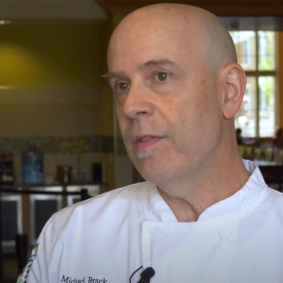 A man dressed in a white chef's coat speaks to the camera in a dining hall kitchen.