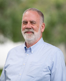 A man in a light blue dress shirt smiles at the camera while standing outdoors.