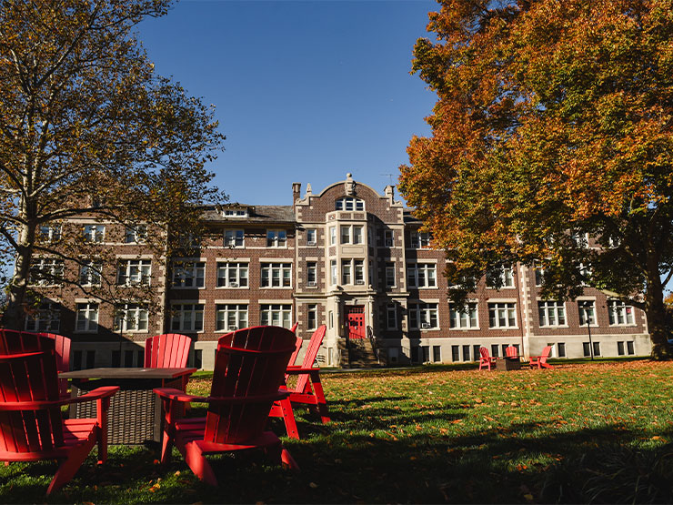 A fire pit and red Adirondack chairs sit on the green lawn in front of Brown Hall, a brown brick residence hall with a red door, on a sunny autumn day.