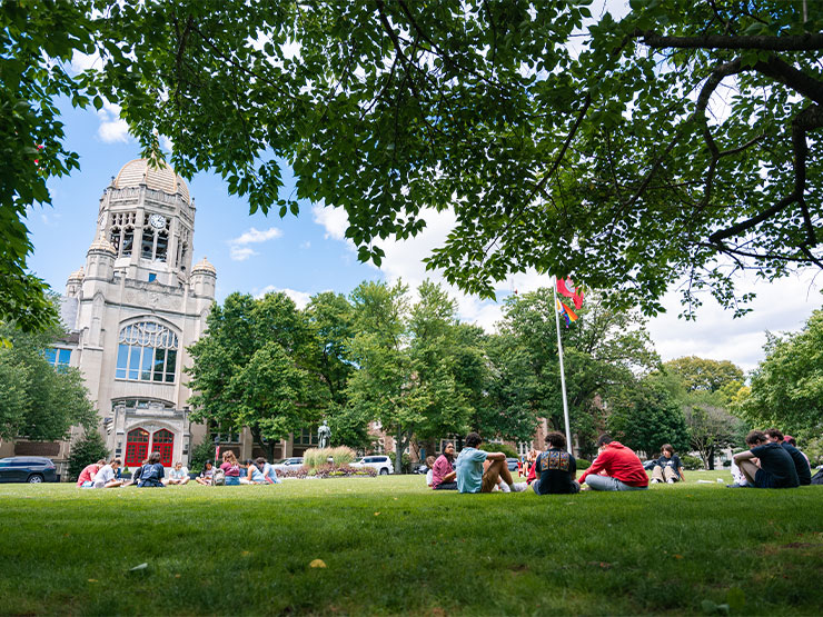 College students sit in groups on the lawn of Muhlenberg College, with the Haas Center College Center clocktower in the distance against a blue sky.