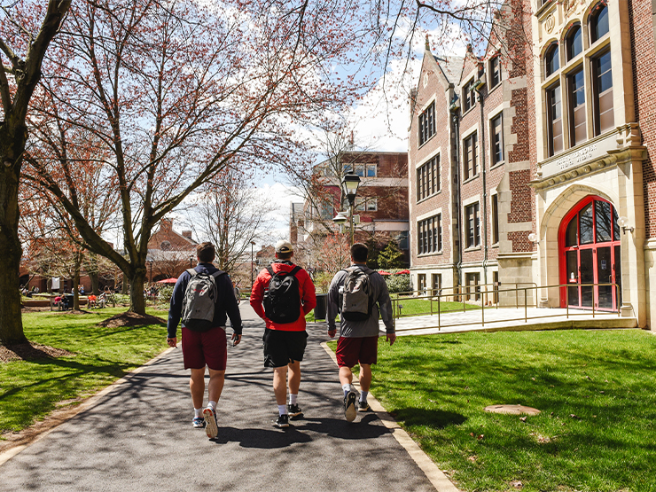 Three students wearing backpacks walk through a college campus, away from the camera.