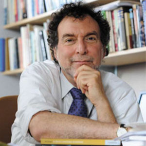 A closeup image of an adult in an office surrounded by books, chin resting in hand.