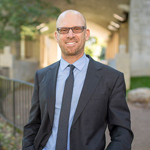 A man in a dark gray and blue suit stands outside, smiling.