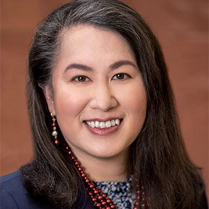 A woman wearing a dark blue suit and red beaded necklace smiles at the camera in a professional headshot.